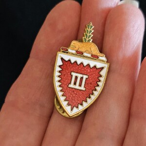 Może przedstawiać: Złota metalowa przypinka w kształcie tarczy z teksturowanym czerwonym środkiem i białą obwódką. Na tarczy widnieje rzymska cyfra III. Nad tarczą znajduje się mała złota figurka zwierzęcia oraz zielony element przypominający liść.