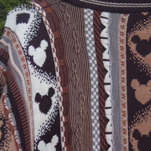 Peut inclure: Gros plan sur un pull multicolore avec un motif de silhouettes de Mickey Mouse. Le pull pr&eacute;sente un m&eacute;lange de rayures verticales marron, beige, blanc et gris. Le tricot a un aspect textur&eacute;.