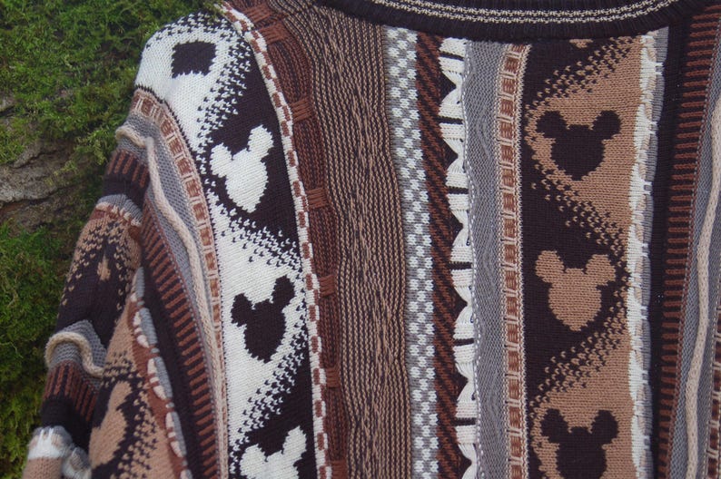Peut inclure: Gros plan sur un pull multicolore avec un motif de silhouettes de Mickey Mouse. Le pull pr&eacute;sente un m&eacute;lange de rayures verticales marron, beige, blanc et gris. Le tricot a un aspect textur&eacute;.