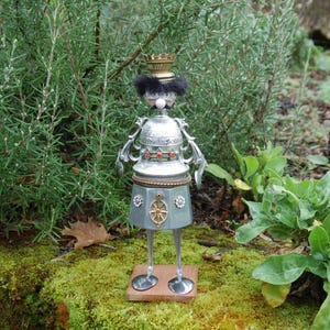 Puede incluir: Una escultura de metal caprichosa de una figura de rey. La figura tiene una corona, un bigote y una base en forma de falda. La escultura está hecha de varios objetos de metal y se apoya en una base de madera.