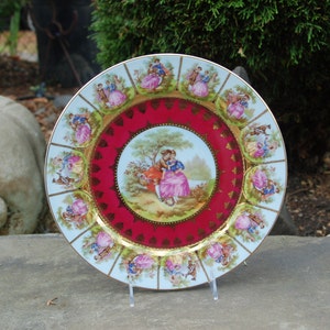Peut inclure: Une assiette en porcelaine blanche avec un bord rouge et dor&eacute;. Le centre de l'assiette pr&eacute;sente une sc&egrave;ne romantique avec deux personnages dans un jardin. Le bord est d&eacute;cor&eacute; d'un motif r&eacute;p&eacute;titif de personnages dans diverses poses.