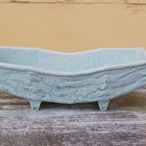 Puede incluir: Un macetero de cerámica azul claro con una superficie texturizada y tres patas. El macetero tiene forma de barco y una abertura rectangular.