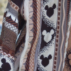 Peut inclure: Gros plan sur un pull tricot&eacute; avec un motif r&eacute;p&eacute;t&eacute; de silhouettes de Mickey Mouse en marron et blanc. Le pull pr&eacute;sente divers panneaux textur&eacute;s dans des tons de marron, beige et gris, cr&eacute;ant un aspect douillet et textur&eacute;.