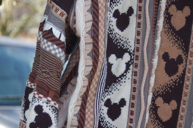 Peut inclure: Gros plan sur un pull tricot&eacute; avec un motif r&eacute;p&eacute;t&eacute; de silhouettes de Mickey Mouse en marron et blanc. Le pull pr&eacute;sente divers panneaux textur&eacute;s dans des tons de marron, beige et gris, cr&eacute;ant un aspect douillet et textur&eacute;.