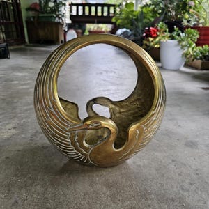 May include: A circular brass vessel featuring a sculpted crane with its wings extended upward to form the top of the frame. The metal has a polished gold-toned finish with recessed areas that show a darker, aged patina along the wing feathers.
