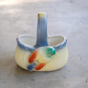 May include: A small, vintage ceramic basket with a handle. The basket is cream-colored with vertical ridges and a blue rim. A colorful bird figurine with red, yellow, and green accents is attached to the side.
