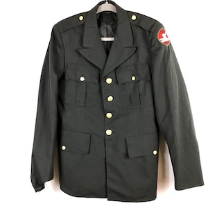May include: Dark green military jacket with gold-colored buttons and a red and white emblem on the upper left sleeve. The jacket has four front pockets and epaulets on the shoulders. The jacket is hanging on a white hanger.