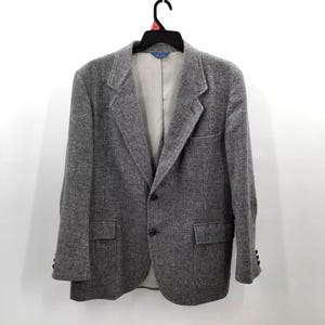 May include: A gray tweed blazer with a two-button closure, two front pockets, and buttoned cuffs. The jacket is hanging on a black hanger against a white background. The interior lining is a light beige color.