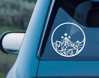 Könnte beinhalten: Weißer geometrischer Berg- und Sternaufkleber auf einer Autoscheibe. Das Design zeigt eine Bergkette mit drei Sternen in einem kreisförmigen Rahmen. Das Auto ist türkis.