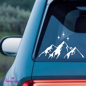 Könnte beinhalten: Weißer Bergketten- und Sternaufkleber auf einer Autoscheibe. Der Aufkleber zeigt eine stilisierte Bergkette mit drei Sternen über den Gipfeln. Der Aufkleber ist auf der Heckscheibe eines blauen Fahrzeugs angebracht.