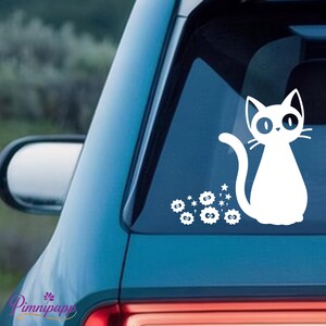 May include: White cat decal with a whimsical design, featuring a stylized cat with large eyes and a curved tail. The decal includes star and flower accents. Applied to a car window.