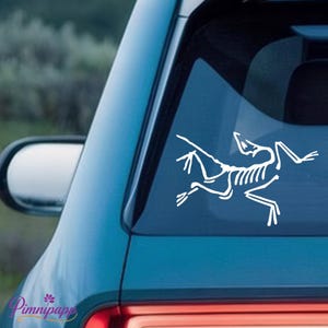 May include: White decal of a stylized skeleton of a creature, possibly a lizard or dinosaur, on a car window. The decal is a minimalist design, featuring the skeletal structure in a running pose. The car is a light blue colour.