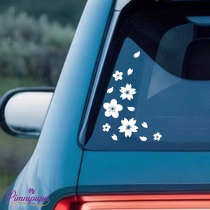 May include: White cherry blossom decals on a teal car window. The decals include various sizes of flowers and falling petals, creating a delicate and artistic design. The car's side mirror is visible.