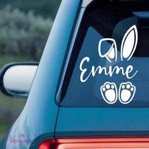 Könnte beinhalten: Weißer Vinyl-Autoaufkleber mit Hasenohren, Pfotenabdrücken und dem Namen "Emme" in Schreibschrift. Der Aufkleber ist für eine Autoscheibe konzipiert und hat ein verspieltes und festliches Thema.