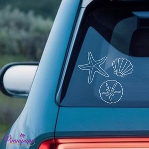 Könnte beinhalten: Weiße Umrissaufkleber eines Seesterns, einer Muschel und eines Sanddollars werden auf einer Autoscheibe angezeigt. Die Aufkleber sind in einer Gruppe angeordnet, mit dem Seestern oben links, der Muschel oben rechts und dem Sanddollar unten.