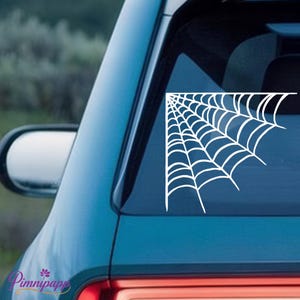 May include: White spiderweb sticker applied to a car window. The spiderweb design is in the corner of the window, with a curved shape. The car is teal, and the background is blurred.