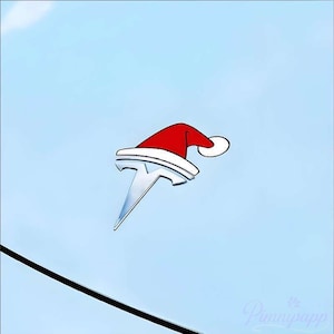 May include: A silver Tesla logo with a red and white Santa hat on a light blue background. The hat has a white trim and a white pom-pom. A black line is visible in the lower left corner.