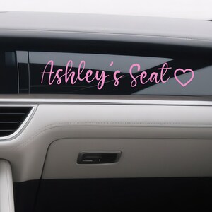 Könnte beinhalten: Rosafarbener Autoaufkleber mit dem Text "Ashley's Seat" und einem Herzsymbol. Der Aufkleber ist auf einer Autoscheibe angebracht, wobei der Innenraum des Fahrzeugs im Hintergrund zu sehen ist.