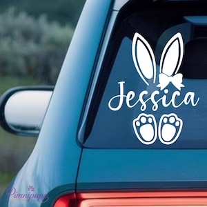 Könnte beinhalten: Weißer Vinyl-Aufkleber mit Hasenohren, einer Schleife, dem Namen "Jessica" und zwei Pfotenabdrücken. Der Aufkleber ist auf einer Autoscheibe angebracht, ideal für Ostern oder Frühling.