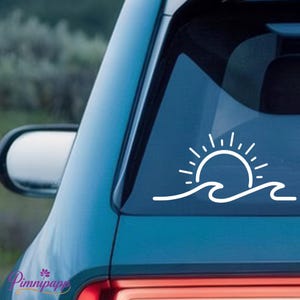May include: A white vinyl decal applied to a car window, featuring a minimalist line art design of a sun rising above a stylized ocean wave. The graphic consists of a semi-circle sun with radiating rays positioned directly over a single continuous wave.