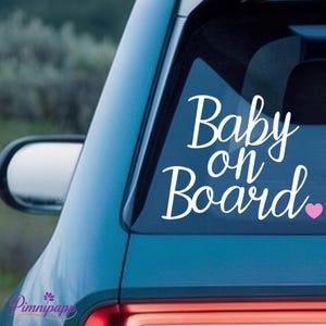 Könnte beinhalten: Weißer Autoaufkleber mit den Worten "Baby on Board" in Schreibschrift und einem kleinen rosa Herz. Der Aufkleber befindet sich auf der Heckscheibe eines tealfarbenen Autos.