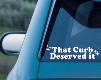 May include: White vinyl car decal with the text "That Curb Deserved it" in a bold, sans-serif font. The decal is applied to the rear window of a teal car, with a side mirror visible.