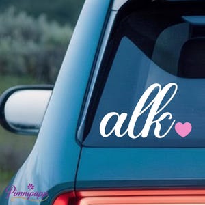 Könnte beinhalten: Weißer Vinyl-Aufkleber mit dem Wort "alk" in Schreibschrift, begleitet von einem kleinen rosa Herz. Der Aufkleber ist auf der Heckscheibe eines türkisfarbenen Autos angebracht, mit verschwommenem Hintergrund.