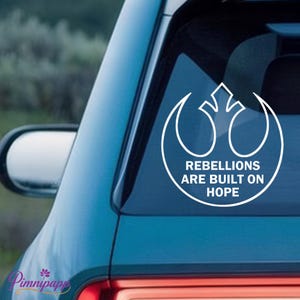 May include: White vinyl decal featuring the Rebel Alliance symbol with the text "Rebellions Are Built On Hope." The decal is applied to the rear window of a teal car. The design is a popular theme.