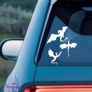 Peut inclure: Autocollants blancs représentant des dragons sur une vitre de voiture. Les décalcomanies présentent des dragons aux ailes déployées et aux corps détaillés. La voiture est de couleur bleu clair, et l'arrière-plan est flou.