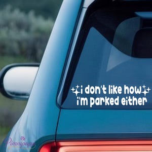 May include: White vinyl car decal with the text "i don't like how i'm parked either" in a playful font. The decal is applied to the rear window of a blue car. Decorative starbursts flank the text.