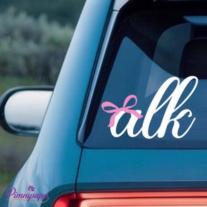 May include: White vinyl decal with the word "alk" in a cursive font, featuring a pink bow. The decal is affixed to the rear window of a teal car. The image is taken outdoors.