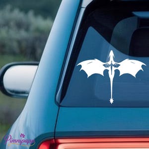 May include: A white vinyl decal of a stylized dragon with large, outstretched wings and a long, pointed tail, applied to the rear window of a blue vehicle. The dragon design features intricate wing details and a star-shaped tip on its tail.