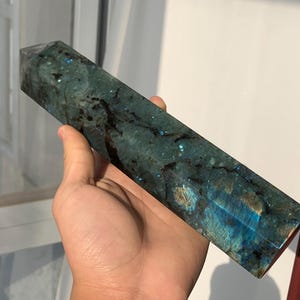 May include: A rectangular, polished labradorite crystal obelisk. The stone has a dark teal base color with black veining and iridescent blue and gold flashes. The obelisk is held in a hand, showcasing its shape and color variations.