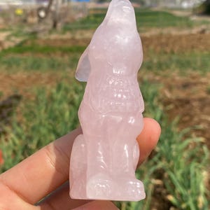 Puede incluir: Una talla de lobo de cuarzo rosa, de pie sobre sus cuatro patas con la cabeza levantada, mirando hacia arriba. La talla es detallada e intrincada, con un acabado liso y pulido.