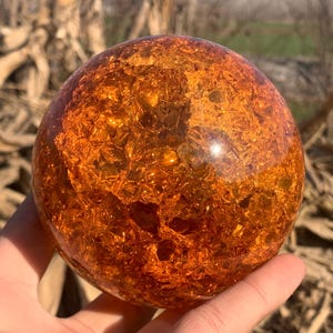 May include: A large, spherical object with a textured, amber-colored surface. The sphere is held in a hand, with the background showing blurred natural elements. The object's color and texture suggest it may be a decorative item or a crystal ball.