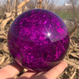 May include: A vibrant, spherical object with a deep purple hue. The surface has a textured, crystalline appearance, catching the light and creating a sparkling effect. The sphere is held in a hand, with a blurred natural background.