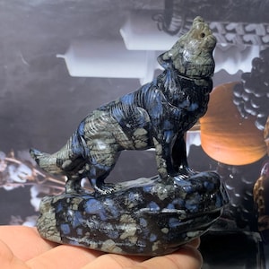 Puede incluir: Una figura de lobo de piedra tallada en tonos azules, grises y negros. El lobo está representado aullando, de pie sobre una base de roca. La escultura tiene texturas detalladas y un acabado pulido, mostrando los patrones naturales de la piedra.