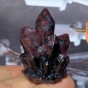 Puede incluir: Un grupo de cristales rojo oscuro con formaciones afiladas y puntiagudas. El cristal tiene una base oscura, casi negra, con vetas rojas y grises más claras. La forma general es irregular y geométrica, parecida a una cordillera en miniatura.