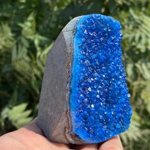 May include: A striking blue crystal geode with a rough, dark gray exterior. The interior is a vibrant, sparkling blue, composed of numerous small crystal formations. The geode is held in a hand, with a blurred green background.