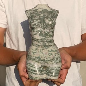 Peut inclure: Une sculpture de torse en pierre sculptée dans les tons de vert et de blanc. La sculpture représente le haut du corps d'une personne, avec des détails précis. La pierre a un aspect naturel et texturé. La sculpture est tenue devant un fond blanc.