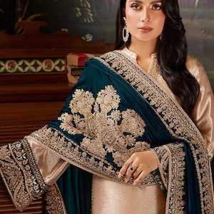 Puede incluir: Una mujer con un vestido color crema y un chal color turquesa con bordados dorados. El chal está drapeado sobre los hombros y los brazos. El vestido tiene un cuello alto y mangas largas. El fondo es una pintura.