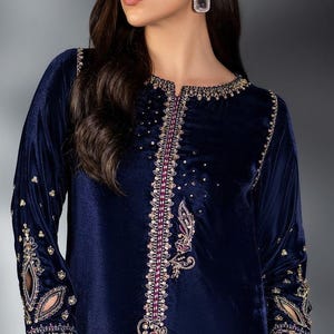 Puede incluir: Un vestido de terciopelo azul marino con intrincados bordados dorados y rosas. El vestido de manga larga presenta adornos en el escote, las mangas y el corpiño. Se sostiene una bufanda rosa transparente.