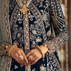 Op de afbeelding: Een close-up van een teal fluwelen kledingstuk met ingewikkelde gouden borduursels. Het ontwerp heeft bloempatronen en versieringen. De mouwen zijn doorschijnend met gouden details. De set is geaccessoireerd met gouden sieraden, waaronder armbanden, ringen en een ketting.