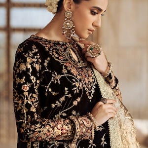 Op de afbeelding: Een donkerbruine fluwelen jurk met ingewikkelde gouden en bloemenborduurwerk. De jurk heeft lange mouwen en een sleutelgat-halslijn. Het model draagt gouden oorbellen, ringen en een armband. Een gouden en crèmekleurige sjaal is gedrapeerd.