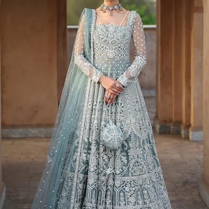 May include: A floor-length, sage green gown featuring intricate silver embroidery, long sheer sleeves, and a square neckline. The ensemble includes a matching sheer dupatta with silver embellishments and a small, round, matching embroidered handbag.