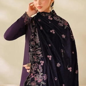Può includere: Una donna indossa un abito viola scuro e uno scialle nero. Lo scialle è adornato da intricati ricami floreali rosa. L'abito ha maniche lunghe con polsini ricamati abbinati. La donna indossa orecchini d'oro e un anello.