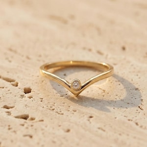 May include: A gold-colored ring with a V-shaped design and a small, round, clear gemstone at the point of the V. The ring is set against a textured, light-colored surface, creating a contrast of textures and colors.