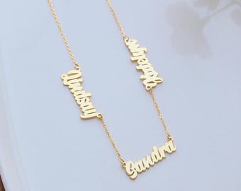 May include: A gold-colored necklace featuring three names: "Lindsay", "Jennifer", and "Sandra" in a cursive font. The names are suspended from delicate chains, creating a personalized jewelry piece. The necklace is displayed on a light-colored surface.