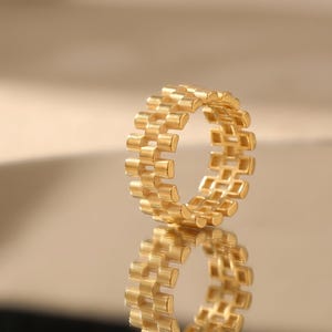 May include: A gold-colored ring with a repeating rectangular pattern. The ring is reflected on a shiny surface. The ring's design is made up of small, connected rectangular blocks, creating a textured appearance.