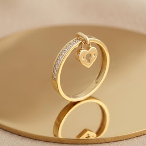 Op de afbeelding: Een gouden ring met een hartvormige hangslotbedel. De ringband is bezet met kleine, heldere stenen. De ring wordt gepresenteerd op een gouden oppervlak, dat de vorm weerspiegelt. De hartvormige bedel heeft een sleutelgatontwerp.